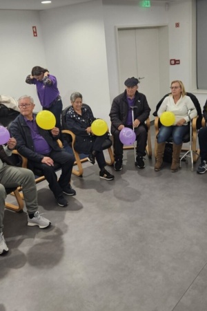 Doze pessoas, de várias idades, sentadas em cadeiras, em meia-lua, metade está de costas e as restantes de perfil, numa sala branca, com uma televisão onde está escrito "Obrigado", ao lado está uma mulher de meia-idade de pé, em frente ao grupo e com um 
